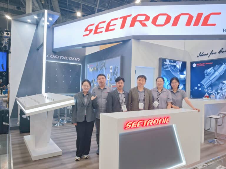 A group stands in front of a "SEETRONIC" booth at a trade show, with promotional material and display items in the background.