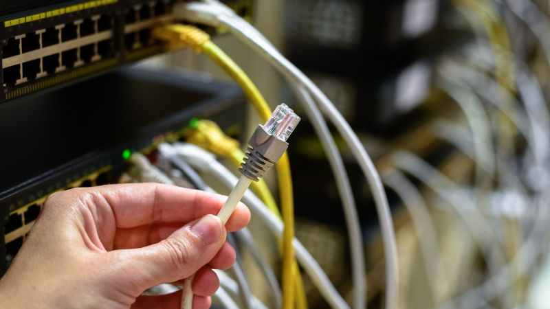 A hand holds an Ethernet cable by a network switch with many connected cables in a server room.