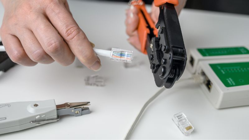 Person crimps an RJ45 connector onto an Ethernet cable with a tester and tools on a white surface.